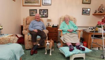 Two elderly people are sitting in arm chairs with a small dog standing in between them