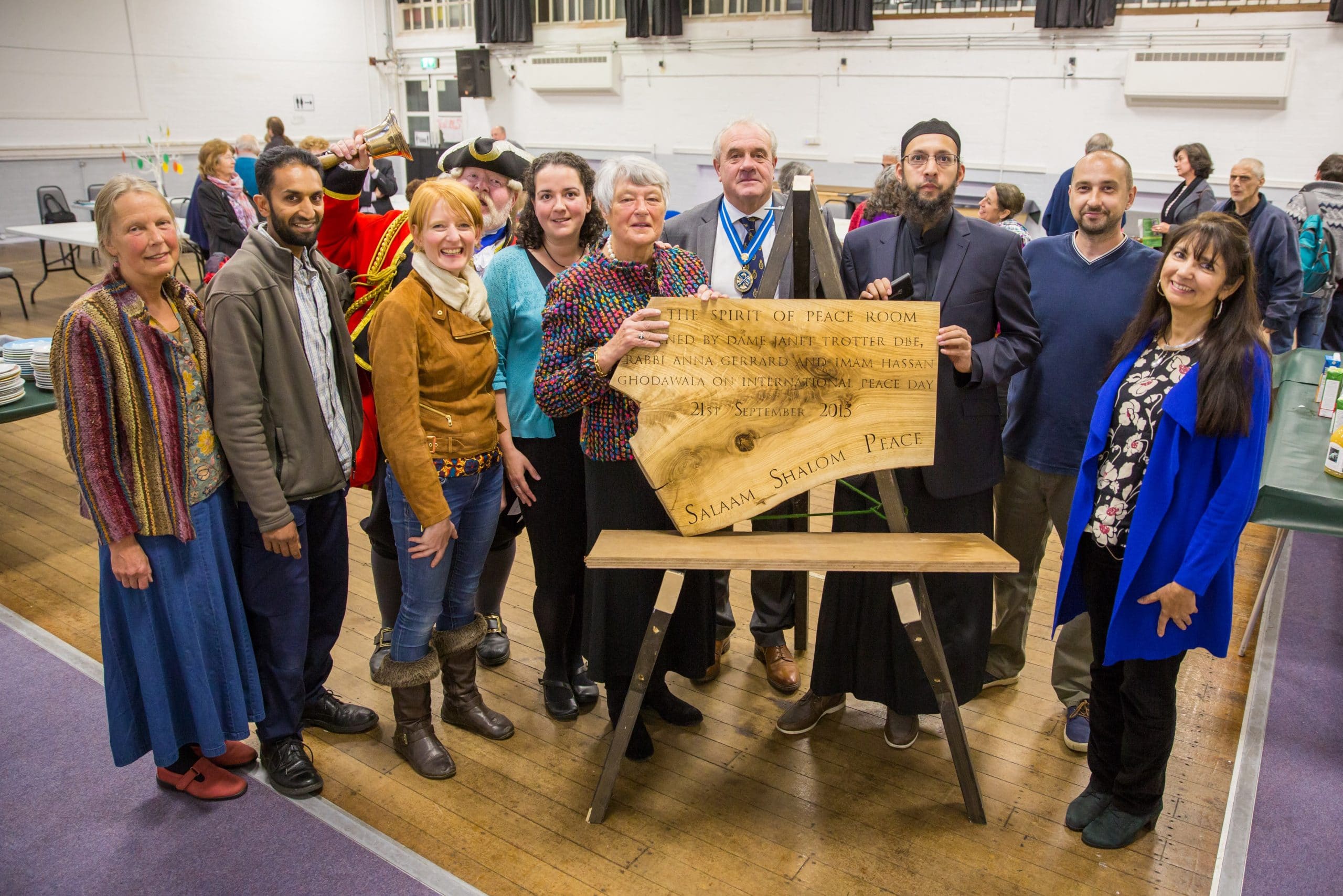 A group of people stands holding a wooden plaque which reads 'Salaam, Shalom, Peace'