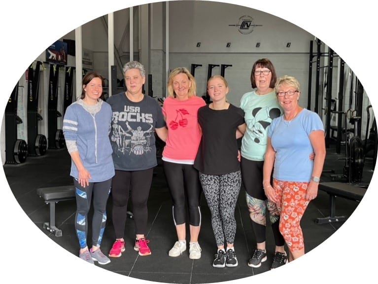 A group of six women stand in front of gymnasium equipment.